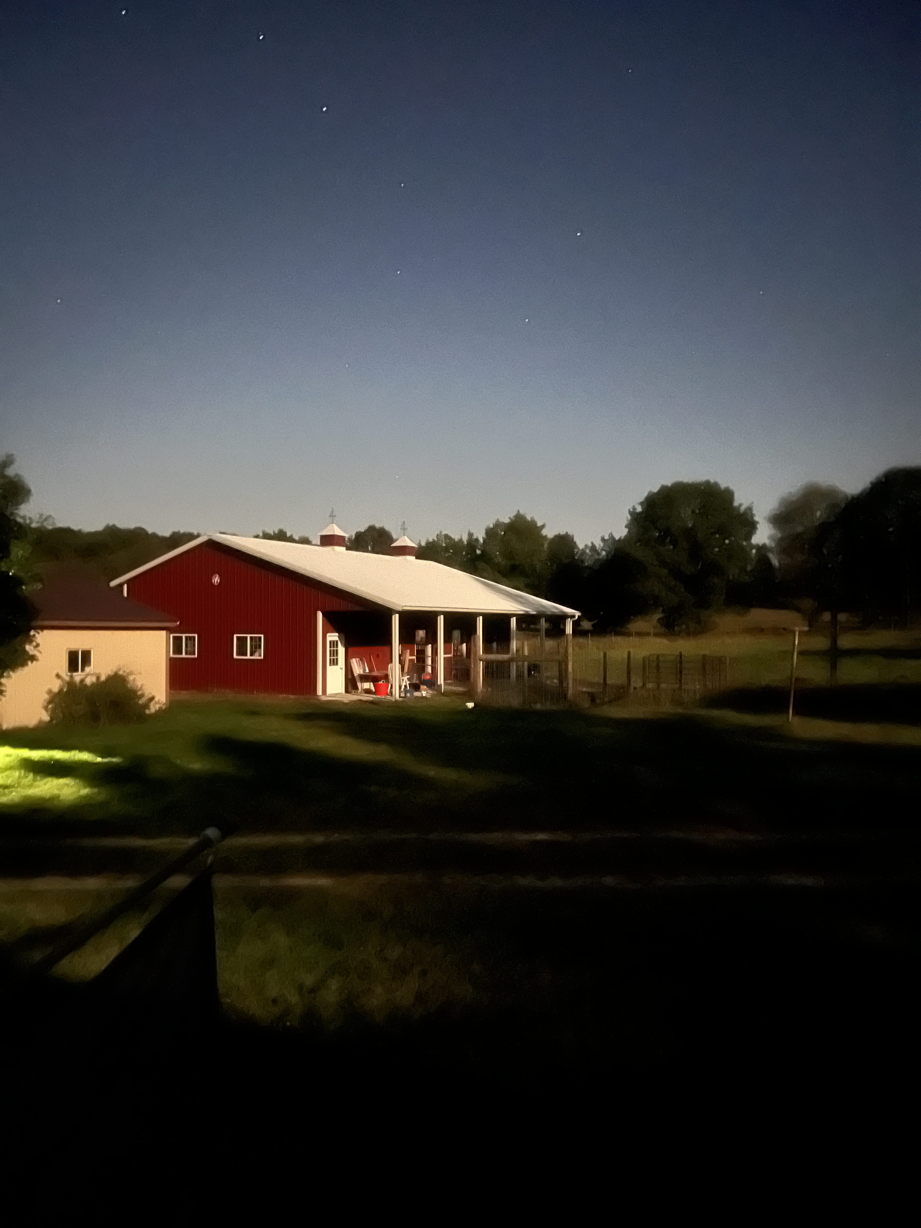 Red barn with a white metal roof lit at night, surrounded by fenced pasture and trees under a starry sky